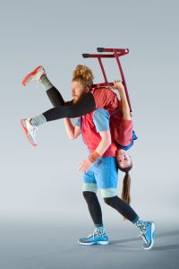 Ein Tänzer trägt eine Tänzerin kopfüber über den Schultern. Ihre Beine baumeln vor seinem Gesicht. Ihr Oberkörper hängt an seinem Rücken herunter. Dabei sitzt sie auf einem Stuhl. Beide tragen auffällige rot-blaue Kleidung.