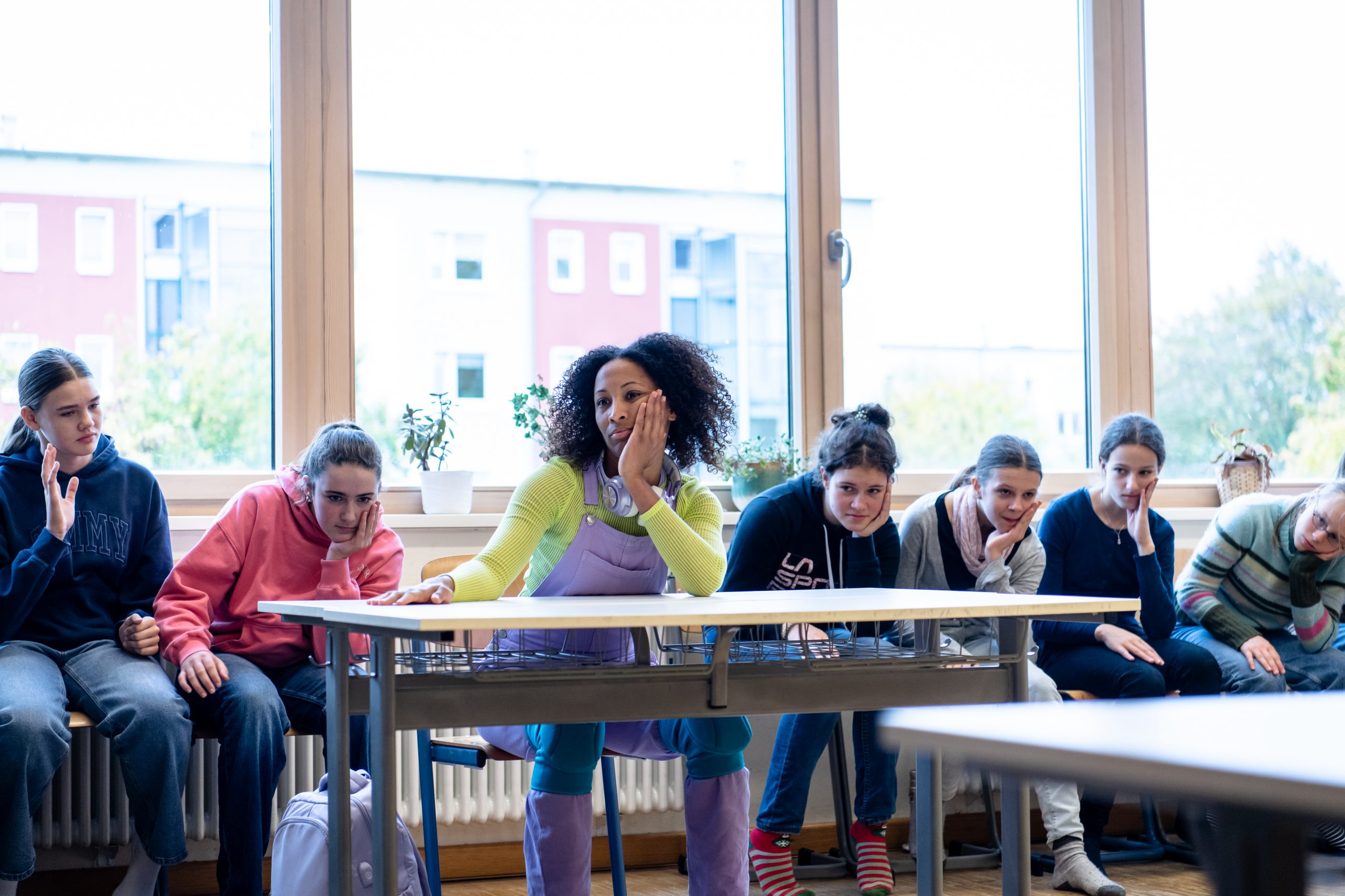 Filimatou Lim sitzt gelangweilt mit aufgestütztem Kopf zwischen den Lernenden in einem Klassenraum am Tisch.