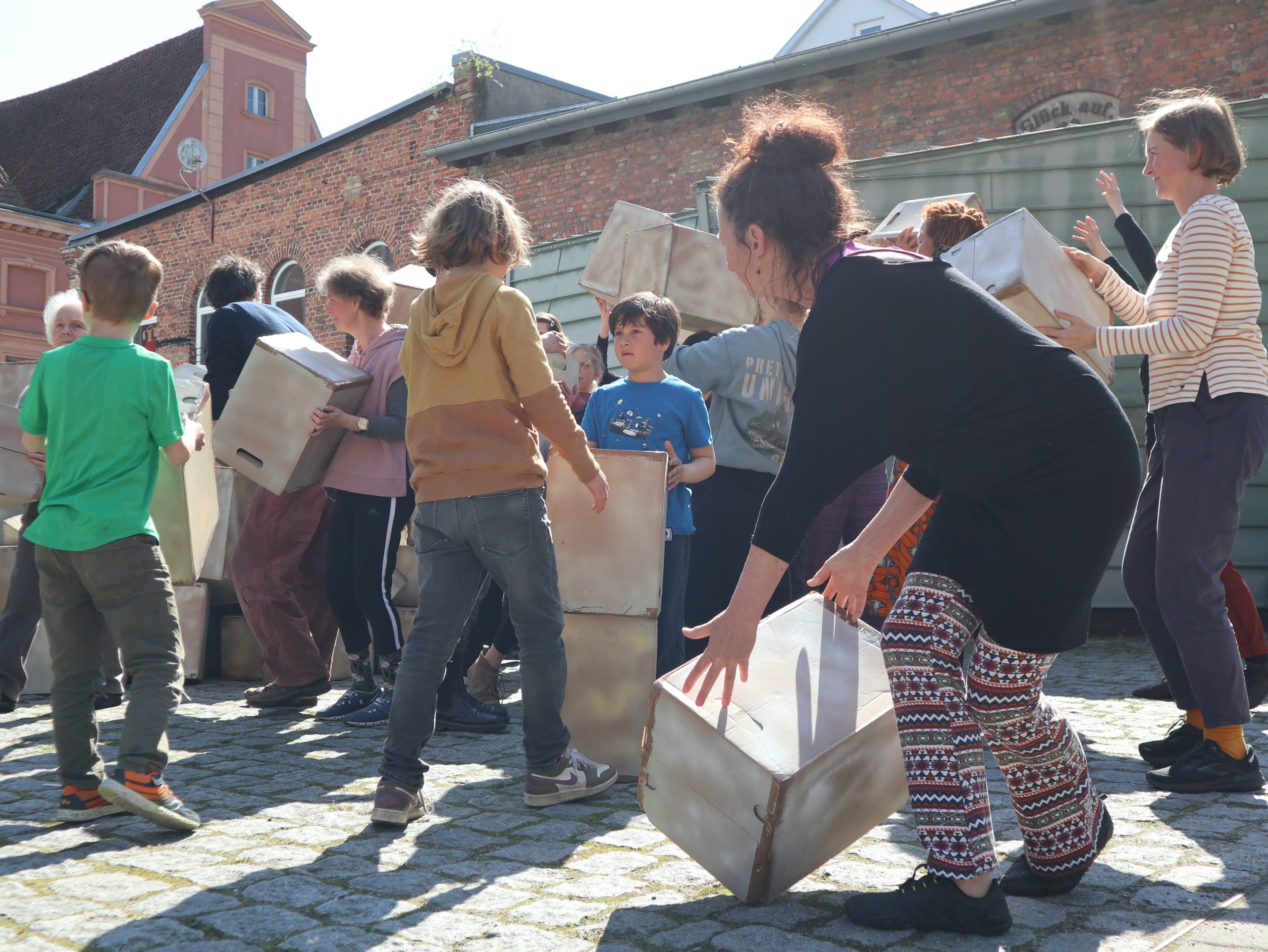 Menschen unterschiedlichen Alters in Alltagskleidung, bewegen sich mit Kisten auf einem öffentlichen Platz.