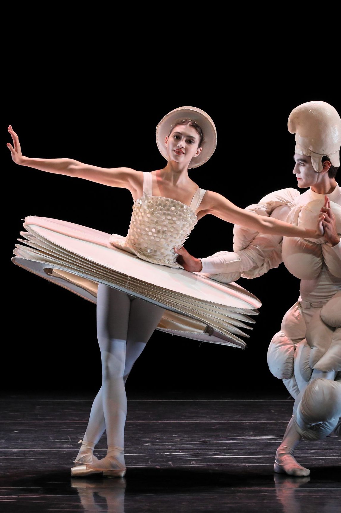 Zwei Tänzer*innen in weißen Kostümen nehmen als Paar Ballettposen ein. Ihre Kostüme haben Auspolsterungen die neue gegenständliche Körperformen kreieren.