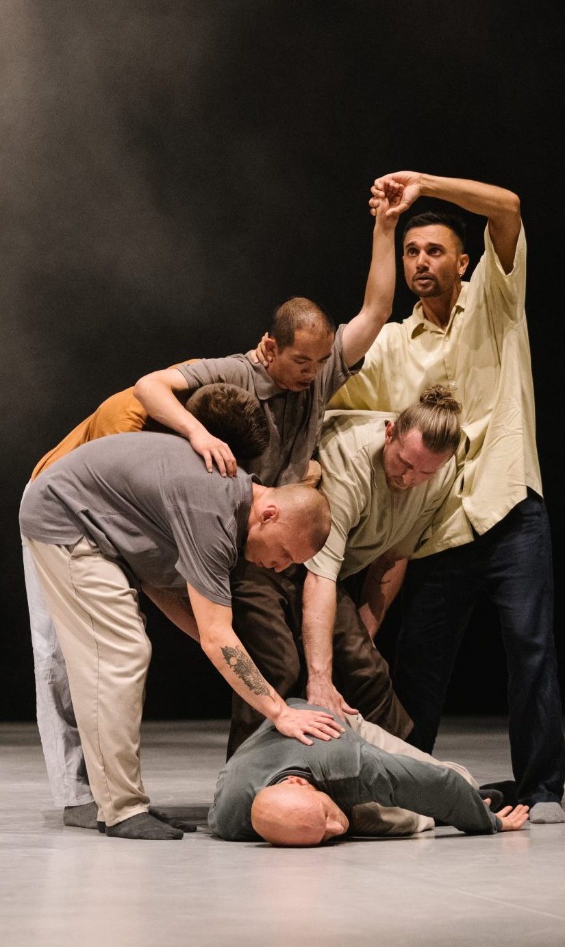 fünf Tänzer in dezenter Alltagskleidung stehen als Gruppe über einen Tänzer gebeugt der mit dem Gesicht nach unten am Boden liegt.